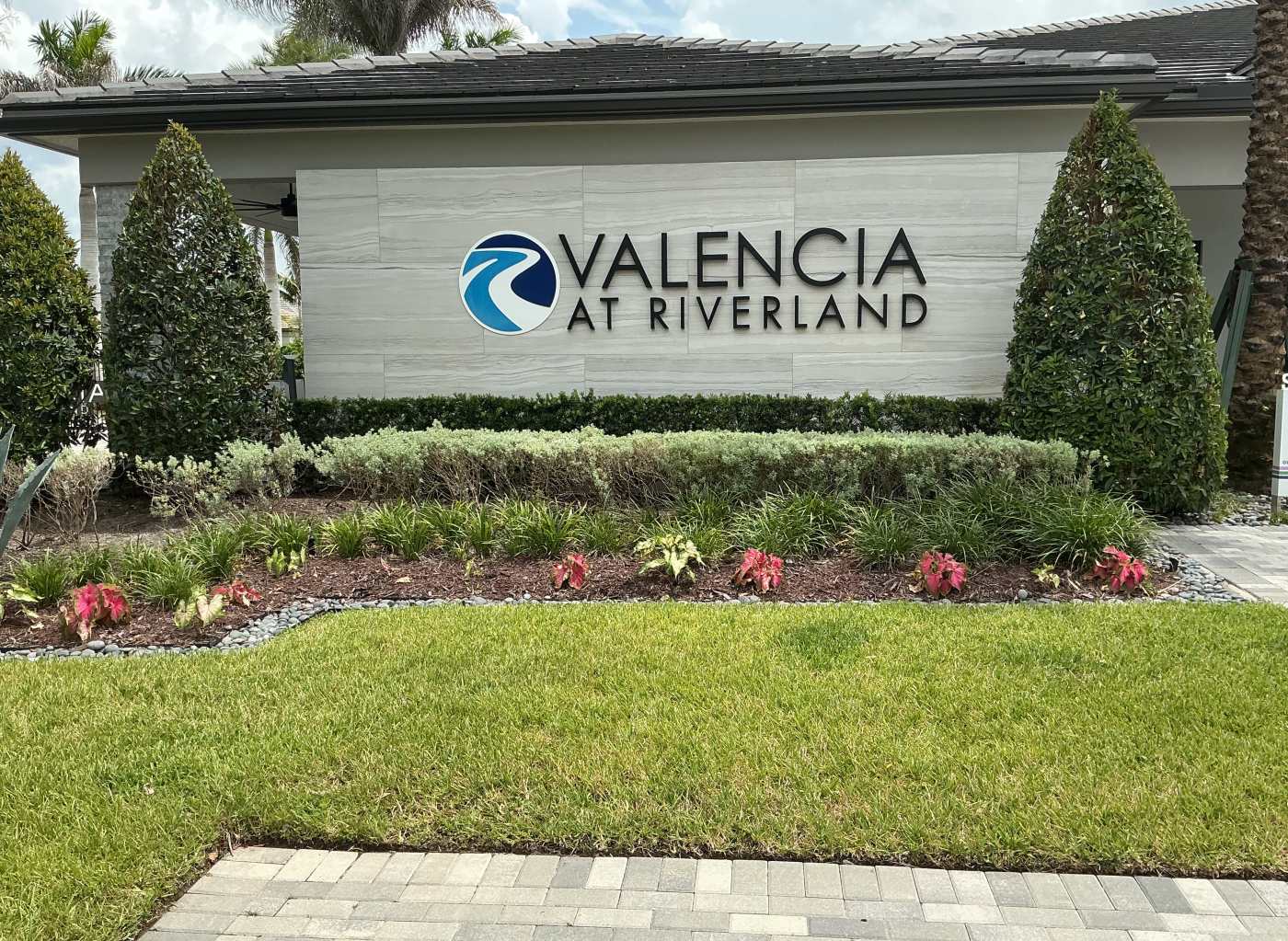 The Valencia at Riverland entrance sign on a stone wall surrounded by landscaped hedges, flowers, and palm trees in Port St. Lucie, Florida